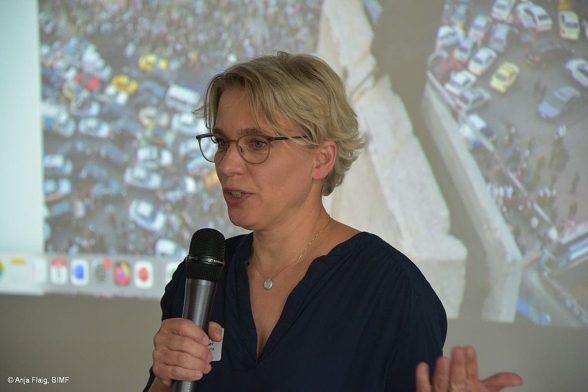 VA Syrien: Referentin Sprechend mit einem Mikrofon in der Hand, im Hintergrund Präsentation